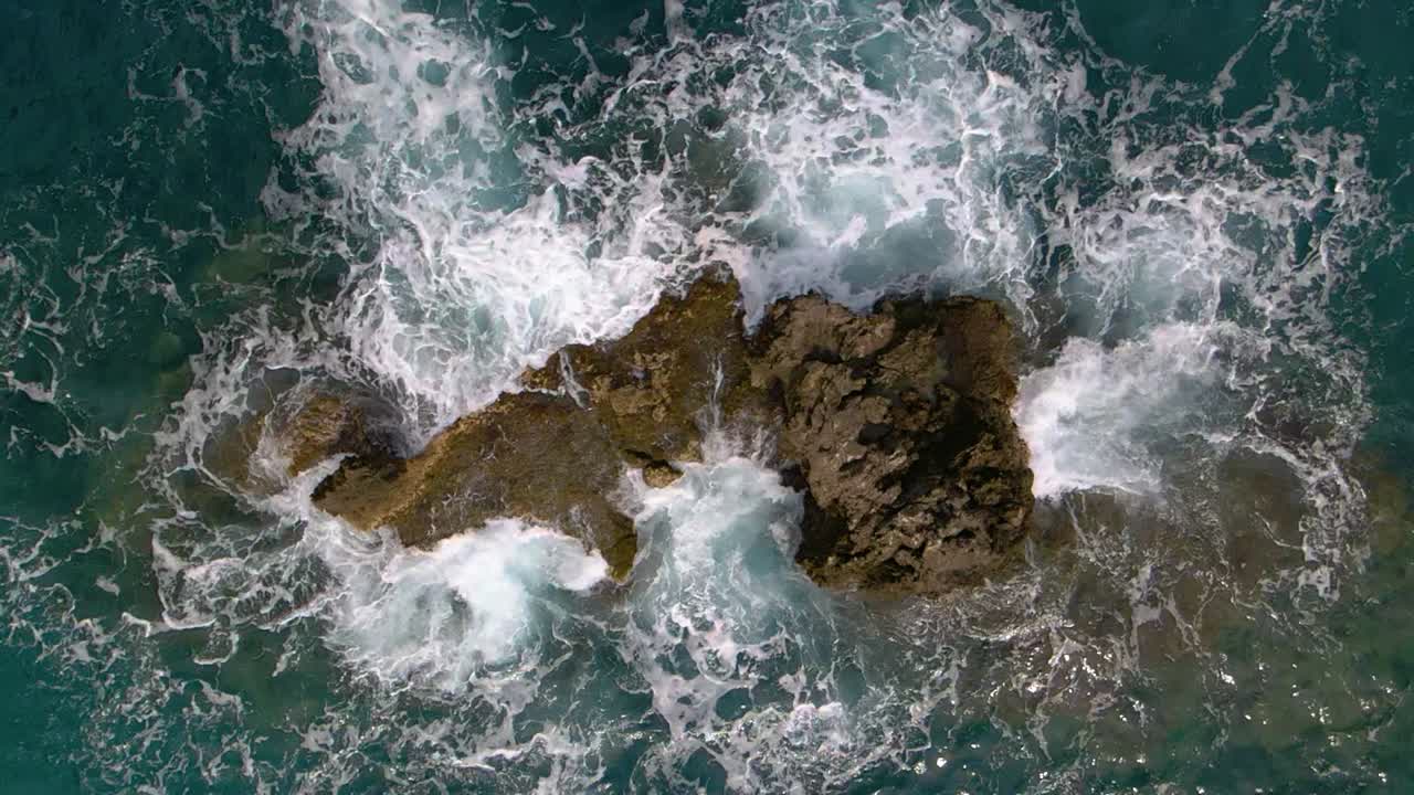 Vista aerea dall'alto dell'acqua del mare che lambisce le rocce, con salita verticale in slow motion a 240fps.