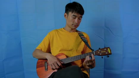 asian man playing a stringed ukulele musical instrument with blue background