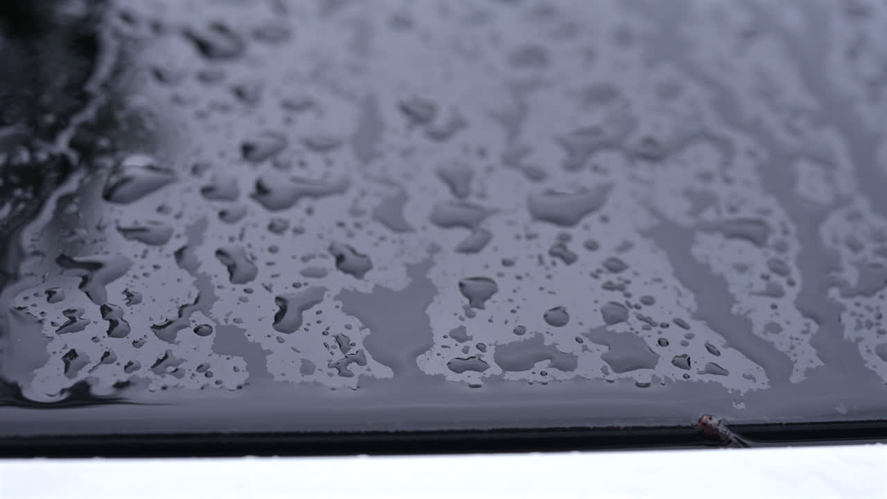 Gros plan de gouttes de pluie recouvrant le toit d’une voiture pendant une averse, avec des reflets flous des bâtiments environnants