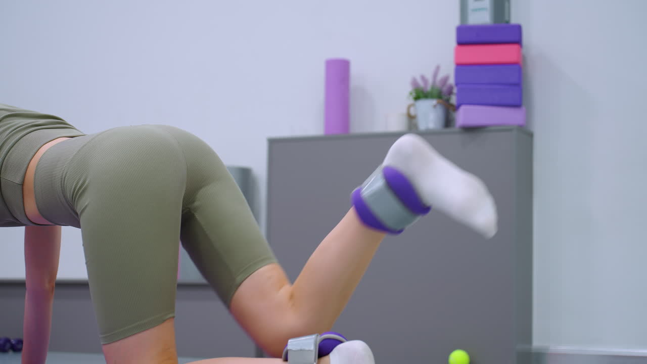 vista parcial de una mujer ejercitando los músculos de las piernas levantando la pierna con un pequeño peso unido, contra un telón de fondo de gimnasio con equipos