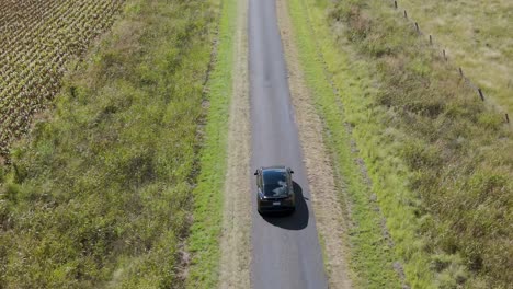 a black car travels along a narrow road flanked by green fields and vegetation.