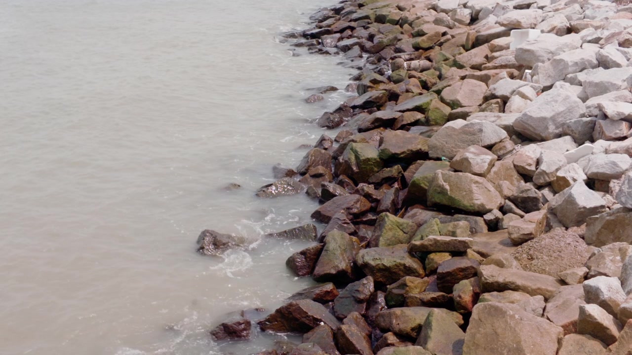 Ripresa statica 4K in slow-motion dall'alto di acqua di mare calma e piccole onde che si infrangono contro una costa rocciosa frangiflutti lungo lo Stretto di Malacca a Melaka, Malesia
