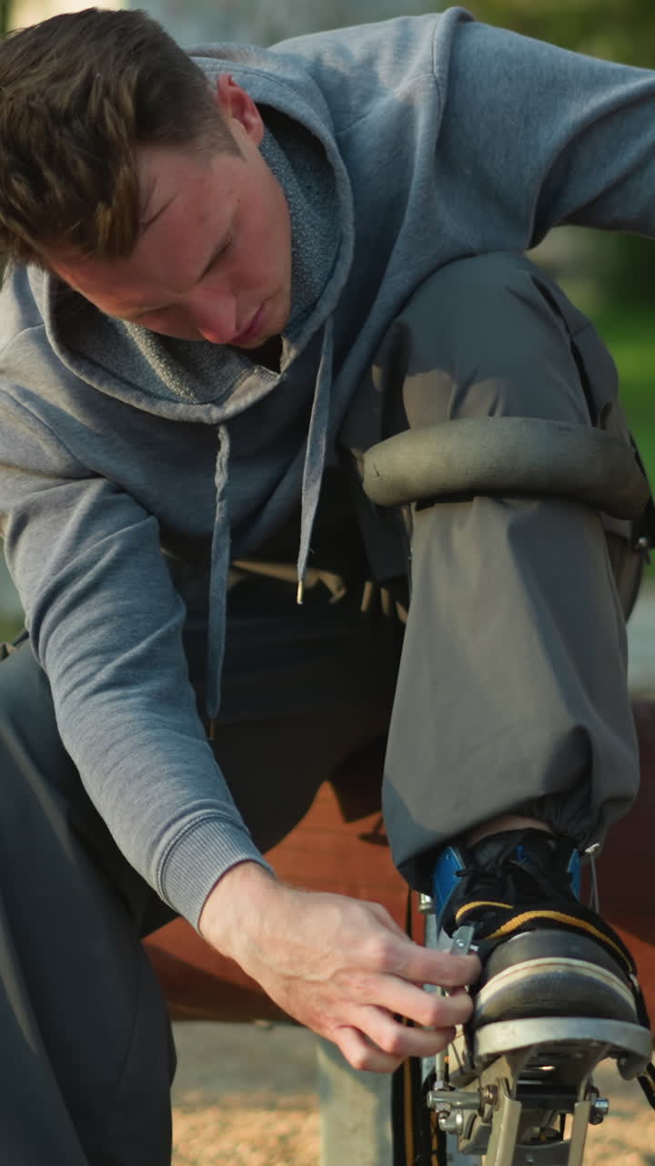 chap sentado en un tronco de madera con capucha gris y pantalones gris oscuro, inclinado para apretar la correa en el zócalo. otro zócalo descansa a su lado en el suelo. entorno natural al aire libre con fondo verde