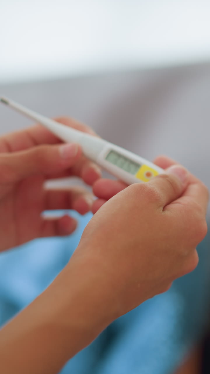 Primo piano di una visuale parziale di personale sanitario che controlla un termometro con una visuale sfocata di una persona sdraiata sullo sfondo, con particolare attenzione al monitoraggio dello stato di salute