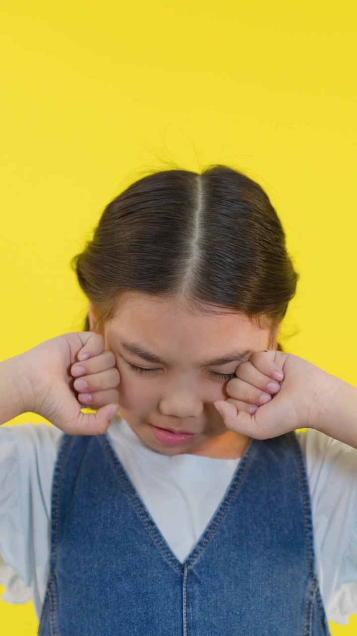 Bambino in abito di jeans si strofina gli occhi con espressione triste, illuminazione brillante da studio, primo piano.