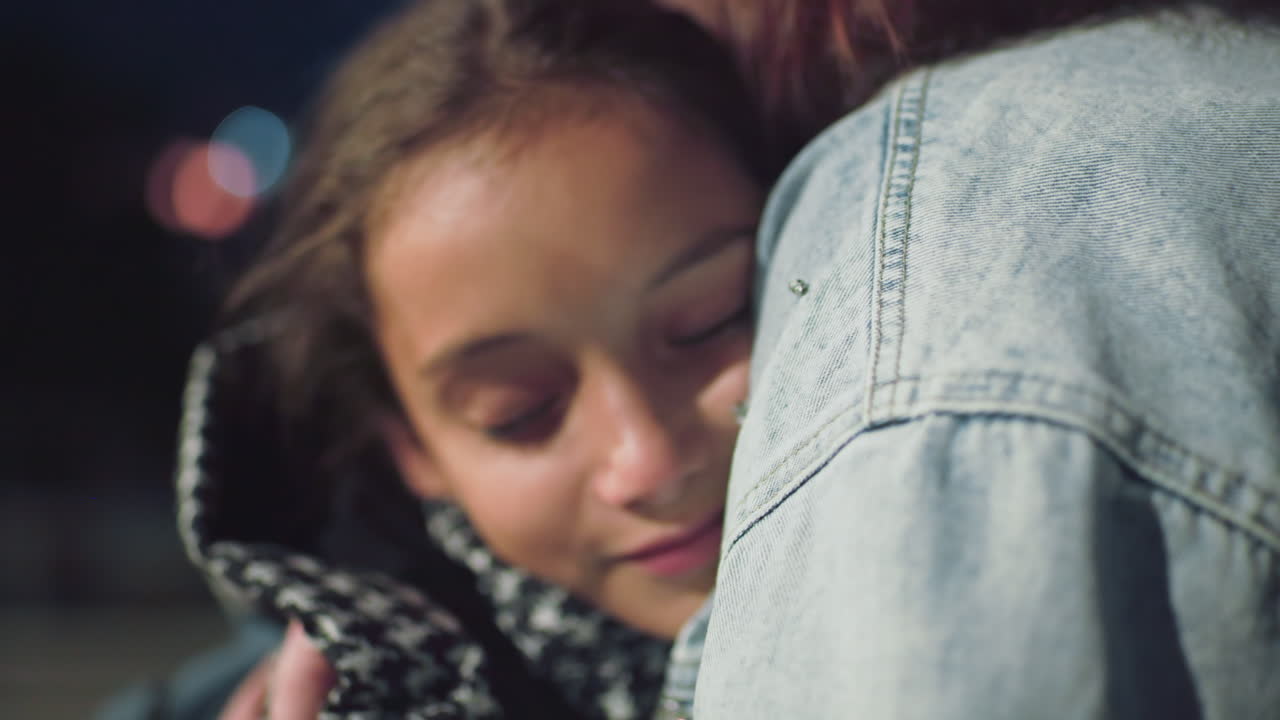 Young girl with soft smile rests in warm hug from woman in denim jacket, eyes looking away thoughtfully, wearing cozy houndstooth coat, showing tenderness, love, emotional bond and gentle connection at night