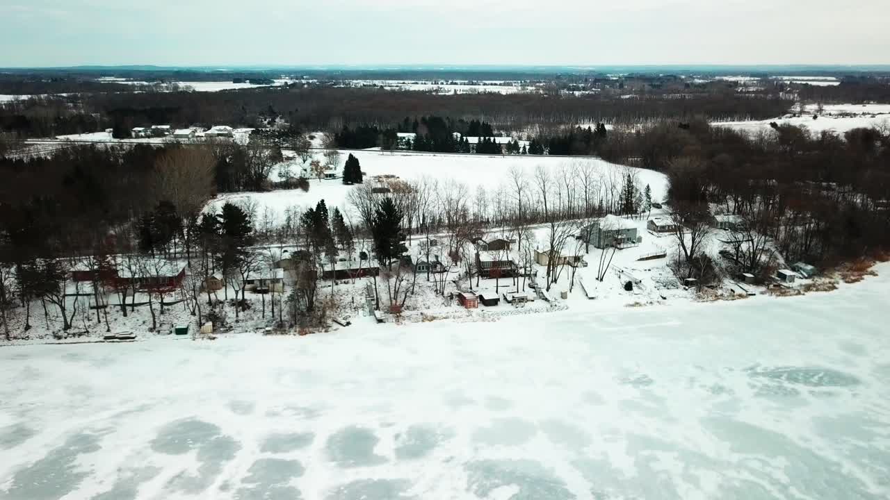 Reverse pull back shot of cabins on a frozen lake in midwestern farmland during winter. 4K