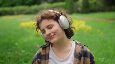 close-up of a happy and cheerful girl with curly hair in a white t-shirt puts on wireless headphones and listens to music on the green grass in the park