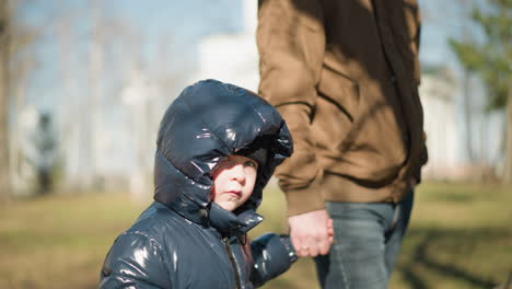 father and son walking hand in hand outdoors, the child, dressed in a shiny black jacket, looks up while they walk together, with a blurred background of trees