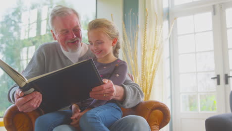 grandfather and granddaughter looking through photo album in lounge at home together