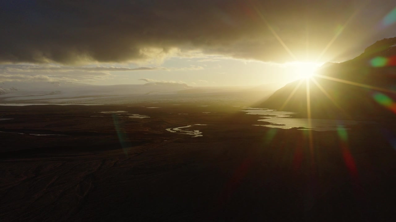 Volcanic wetlands at Herdisarvik, Olfus, South Iceland, extend toward the Atlantic Ocean with winding channels, dark lava terrain and golden sunlight through heavy clouds, drone forward flyover.