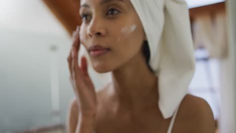 Mixed-race-woman-wearing-towel-on-head-applying-cream-on-her-face