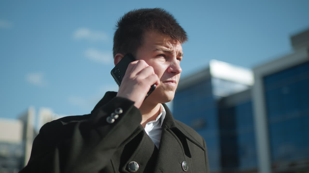 금속 버튼으로 된 검은 코트를 입은 계약자는 야외에서 전화 통화를 하고, 상적으로 보이며, 흐릿한 배경의 현대적인 건물과 함께 밝은 하늘 아래 깊은 숨을 쉬고 있습니다.