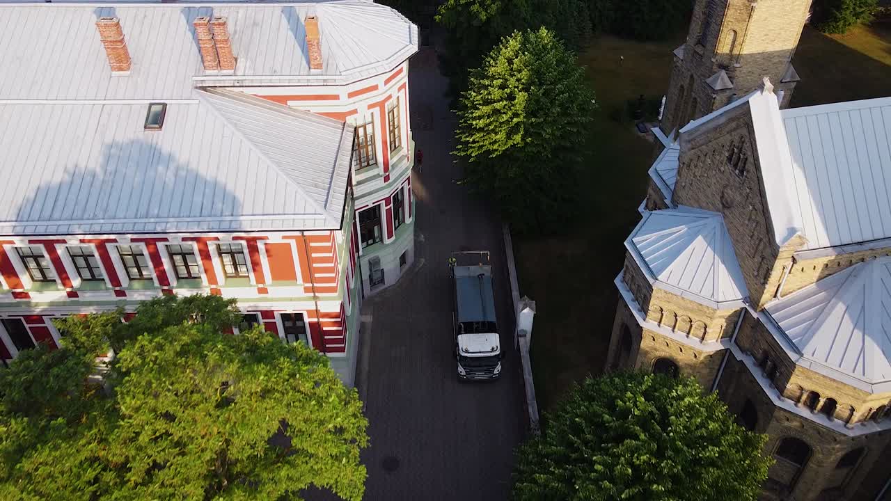 쓰레기를 자동으로 적재한 후 쓰레기 트럭의 공중 전망, 쓰레기 수거통 이동, 일출 후 이른 여름 아침, 푸른 나무, 광각 버드아이 드론 샷이 뒤로 이동