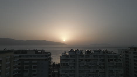 Drone-shot-above-the-city-Vlore-Albania-looking-over-the-buildings-and-harbour-with-ferry-with-the-sea-and-beach-underneath-during-sunset-in-the-evening-with-an-orange-glow-LOG