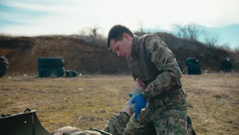 a confident young man in army camouflage and blue medical gloves puts a thick fabric bandage on the hand of his teammate during combat operations in the steppe territory