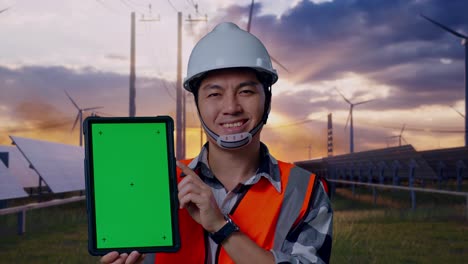 close up of asian male engineer with safety helmet smiling and showing green screen tablet to the camera while standing with solar panel and wind turbines