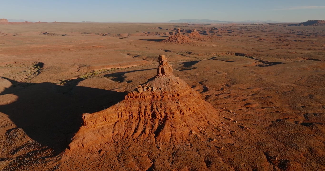 Drone tracks left over a glowing layered sandstone butte at Valley of the Gods, Utah, with Rooster Butte and the sweeping red desert floor bathed in warm evening light