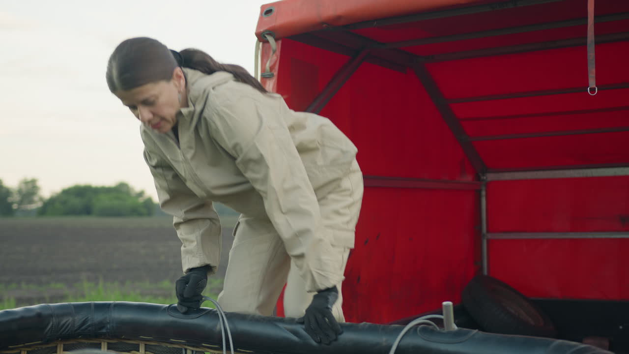 Focused woman in light jumpsuit and gloves connects wire into balloon basket frame while standing beside red truck, preparing for hot air balloon launch in calm rural setting under early morning sky