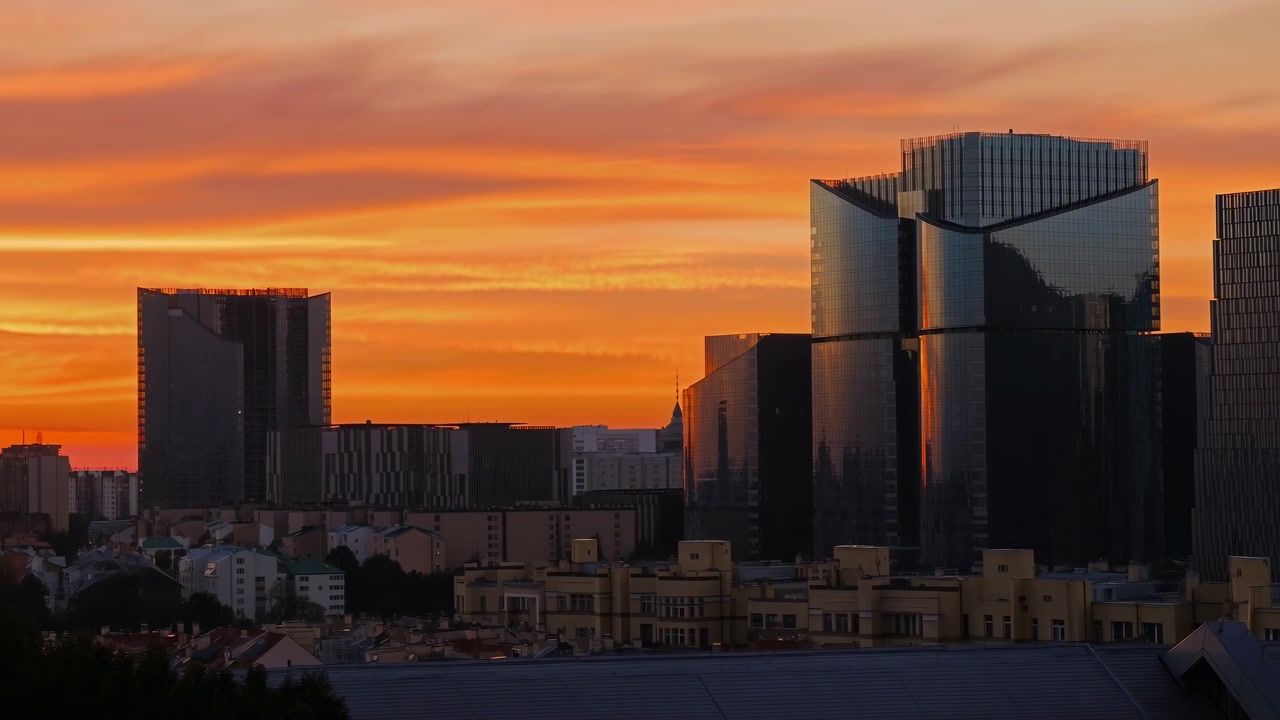 Ampla paisagem urbana ao entardecer mostrando arranha-céus modernos espelhados à direita acima de edifícios baixos em primeiro plano sob um céu laranja vívido.