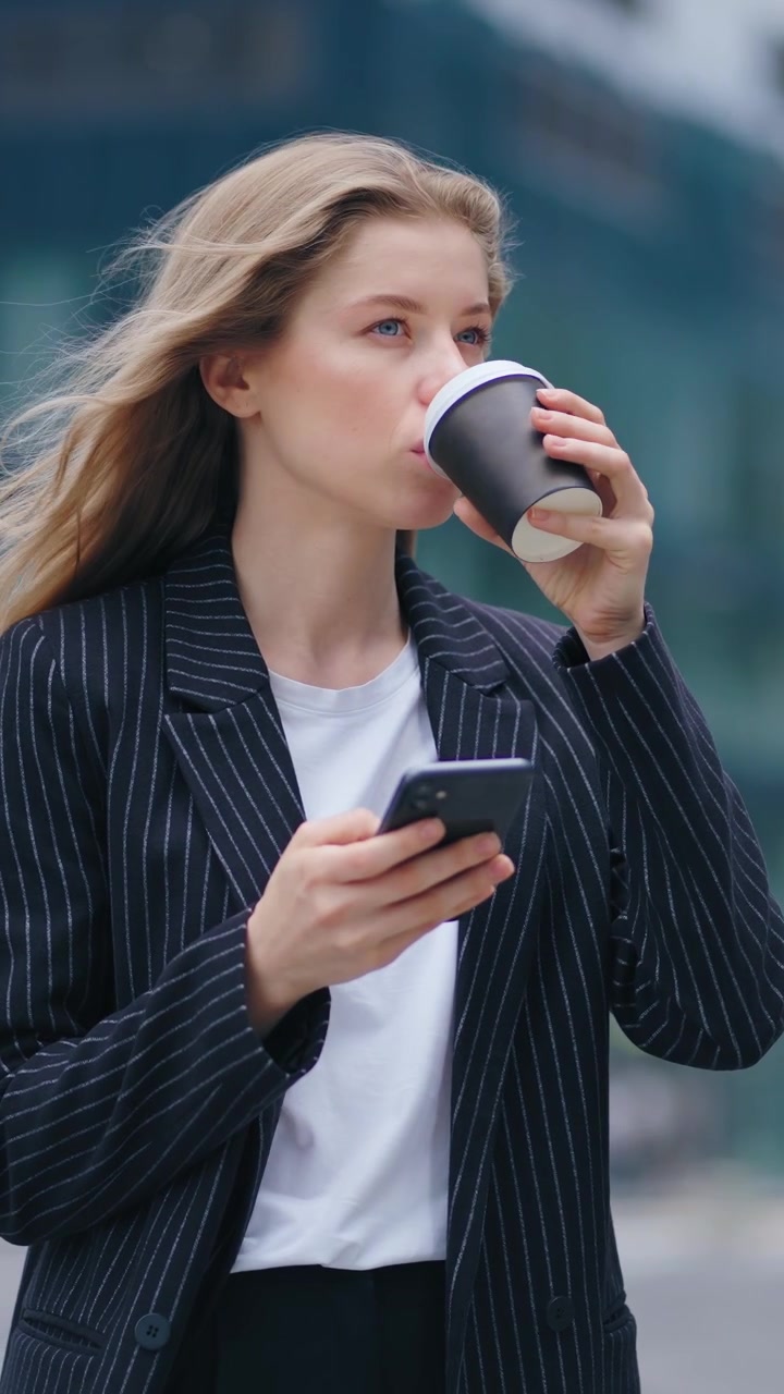 Ung forretningskvinde bruger smartphone og drikker kaffe udendørs.