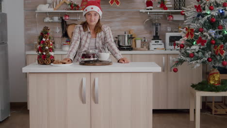 retrato de una chica con sombrero de santa mirando a la cámara de pie en la mesa en la cocina decorada con navidad