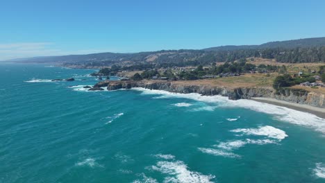 High-altitude drone view over the ocean showing California coastline, sandy beach, rugged cliffs, and houses behind the shore under a clear blue sky