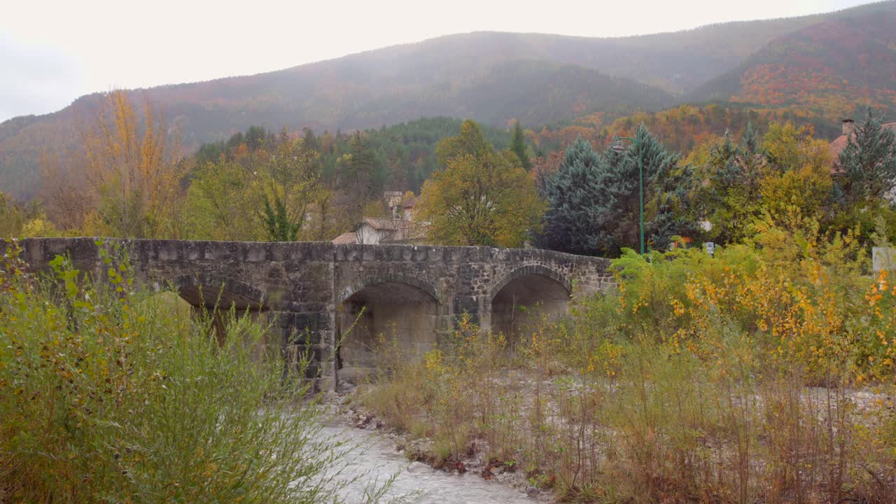 Un sereno ponte di pietra attraversa un ruscello placido, circondato da vivaci colori autunnali nella regione della Provenza. Ideale per promozioni turistiche o documentari sulla natura.
