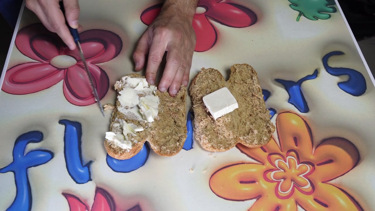 Top down close-up view of male hands using a knife to spread block butter onto a sliced brown wheat bun. Preparing a simple homemade breakfast sandwich on a colorful floral table indoors.