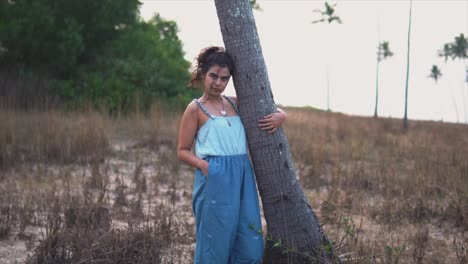 una joven con un vestido azul sostiene un árbol en una playa cubierta de maleza y mira la cámara