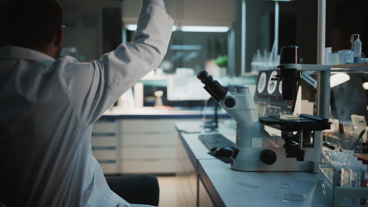 Surprized Male Reseach Scientist Makes an Important Discovery While Researching Virus Vaccine Samples Under the Microscope. He Calls Colleagues and Shares His Success with Fellow Bioengineers.