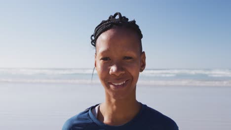 Mujer-Afroamericana-Mirando-La-Cámara-Y-Sonriendo-En-La-Playa-Y-El-Fondo-Del-Cielo-Azul