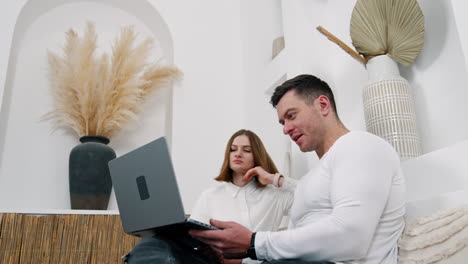 man and woman sitting on the white sofa are focused on laptop. couple working remote from their home. low angle view.