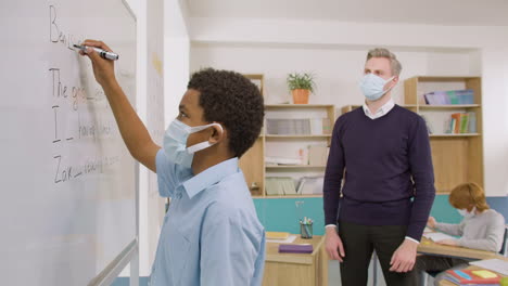 side view of student wearing face mask writing a sentence on blackboard in english classroom while teacher watching him 1