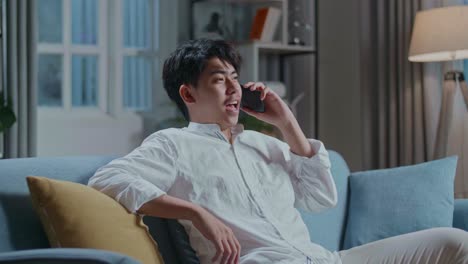 smiling asian man answering the call on smartphone while sitting on sofa in the living room