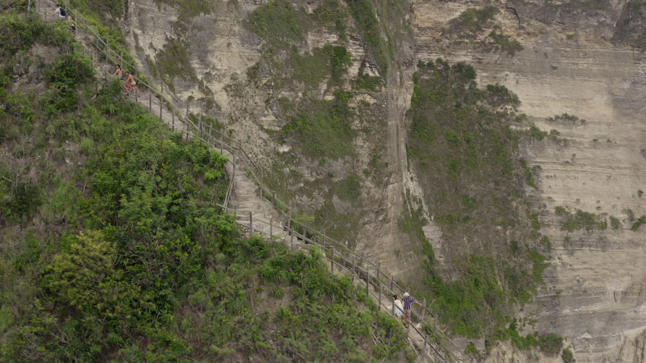 Tourists walking along a steep ridge with a pathway to Kelingking Beach with huge cliff wall in the background, Nusa Penida, Bali, Indonesia. Aerial shot