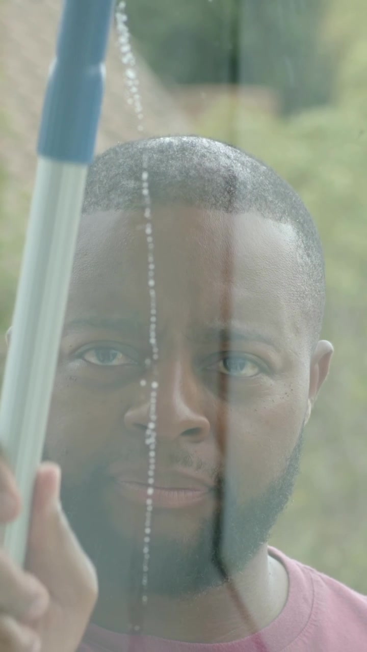 Vertical video: Inspecting glass streaks, man in maroon shirt reaching blue squeegee, wiping window