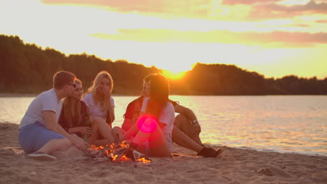 la joven rubia está contando una historia a sus amigas alrededor de una hoguera en la playa. están bebiendo cerveza al atardecer y disfrutando de la noche de verano en la costa del lago.