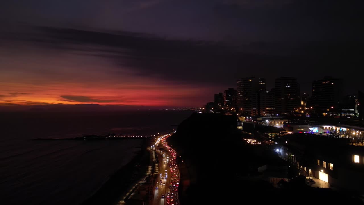 una toma aérea muestra el final de una puesta de sol en una ciudad costera de américa latina, donde se puede ver un centro comercial, edificios y una carretera con tráfico de automóviles bajo un barranco