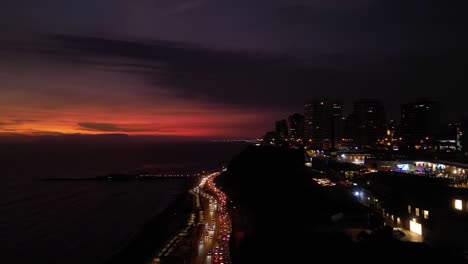 una toma aérea muestra el final de una puesta de sol en una ciudad costera de américa latina, donde se puede ver un centro comercial, edificios y una carretera con tráfico de automóviles bajo un barranco