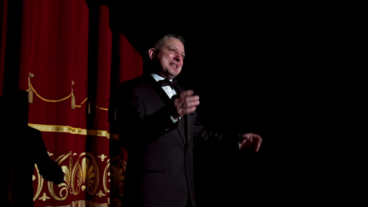 Male host talking on stage. Performer man in black suit standing on dark stage at spotlight and speaking with gestures in the theater. Circling camera.