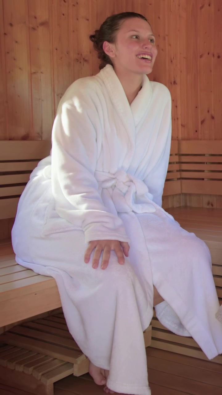 Video verticale: Ospite sorridente della spa che si sistema l'accappatoio bianco su una panca di legno della sauna, rispondendo a una voce.