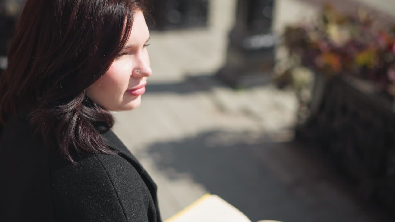 젊은 여성은 갈색 머리카락이 흐르는 머리를 돌리고, 눈에 띄는 코 반지, 도시 환경에서 주변을 둘러보며, 그녀가 우아하게 움직일 때 꽃이 꽃을 피운다.