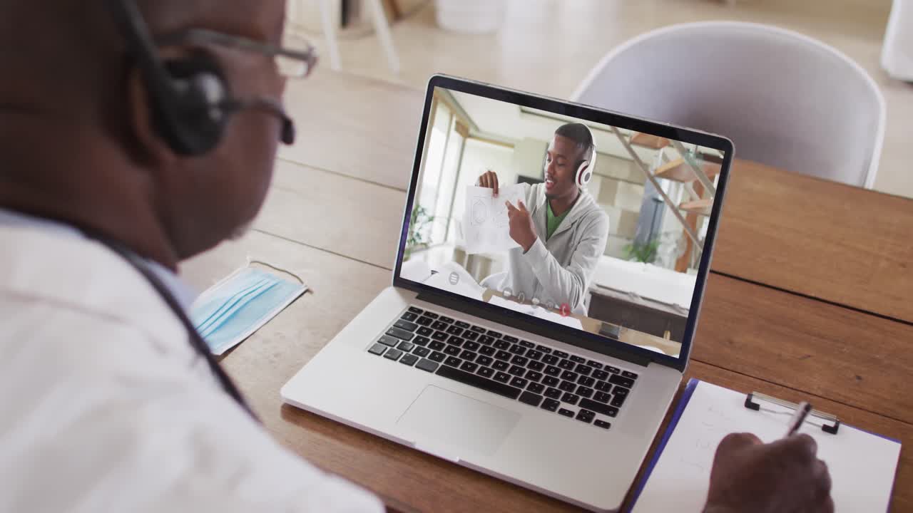 Ein afroamerikanischer Arzt mit Telefon-Headset macht sich Notizen während eines Videoanrufs am Laptop.