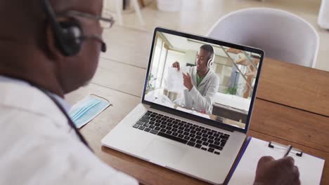 african american male doctor wearing phone headset taking notes while having a video call on laptop