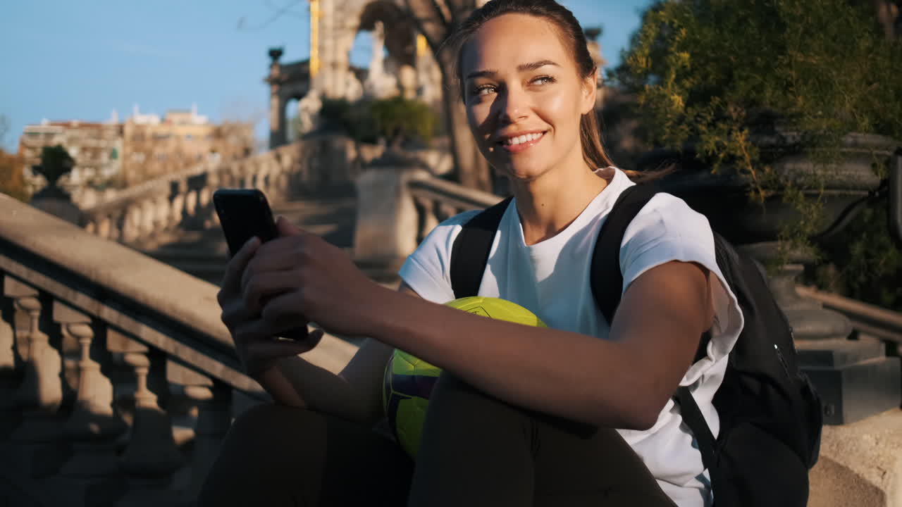 mujer sonriente con teléfono inteligente descansando en las escaleras en el parque de la ciudad después de jugar al fútbol.