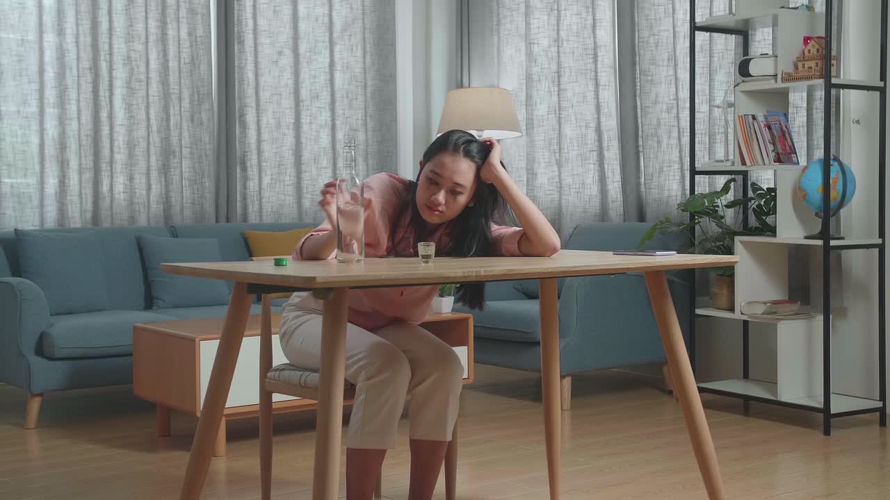 술에 취한, 우울한 아시아 여성이 테이블 위에 스마트폰을 들고 집에서 술을 마시기 전에 크 컵에 보드카를 부었습니다.