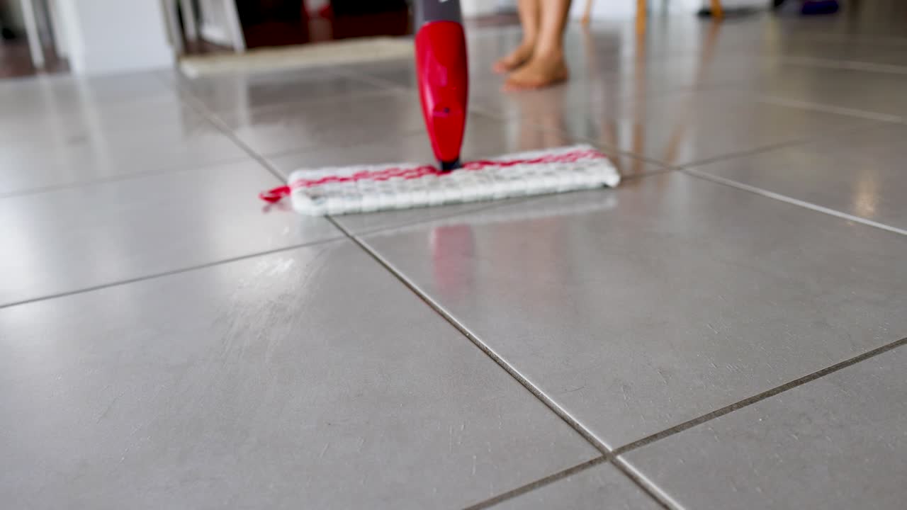 A red spray mop glides over a tiled floor, enhancing cleanliness with water spray. Bright lighting highlights the cleaning process