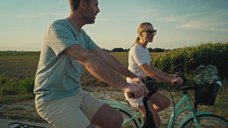 Fröhliches-Kaukasisches-Paar-Mittleren-Alters,-Das-Bei-Sonnenuntergang-Auf-Der-Dorfstraße-Fahrrad-Fährt.