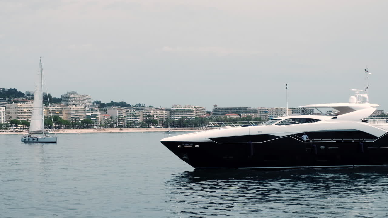 Un elegante yacht in bianco e nero scivola sul mare calmo vicino alla costa della Costa Azzurra, con edifici moderni e barche a vela sullo sfondo.